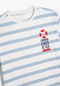 Camiseta blanca con rayas horizontales azul claro. Presenta un gráfico de un salvavidas rojo y blanco y el texto "SALVANDO VIDAS EN EL MAR" en negro.