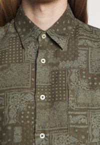 Bruine shirt met een paisleypatroon in lichtblauw. De stof heeft een gestructureerde afwerking, met een klassieke kraag en knopenlijst.