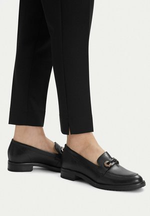 Person trägt schwarze flache Ledermokassins mit Metall-Schnallendetail, kombiniert mit schwarzen, schmal zulaufenden Hosen mit Knöchelschlitzen.