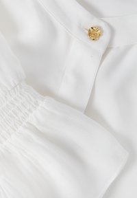 Blouse blanche en tissu texturé, manches froncées et bouton décoratif doré. Surface lisse avec un léger éclat, sans motifs visibles.