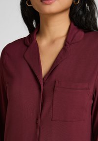 Burgunderrotes, knöpfbares Pyjamaoberteil mit Kragen, versehen mit einer Brusttasche und glatter Stoffstruktur, vor neutralem Hintergrund dargestellt.