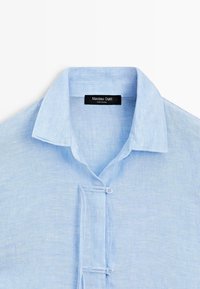 Lichtblauw linnen overhemd met een klassieke kraag en twee geknoopte stoffen knoopsluitingen aan de voorkant.