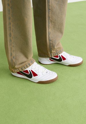 Bijele Nike tenisice s crvenim i crnim akcentima, s teksturiranom površinom i smešnim gumiranim đonom, nošene sa svijetlosmešanim trapericama.
