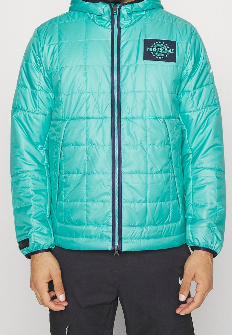 Veste matelassée à capuche de couleur sarcelle avec fermeture éclair foncée, patch "Internazionale" sur la poitrine, portée par une personne en pantalon noir avec logo Nike.