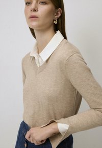 Beige v-hals trui, zachte textuur, aangesloten stijl, gelaagd over een wit overhemd met kraag. De manchetten hebben een sleufdetail. Blauwe spijkerrok zichtbaar.