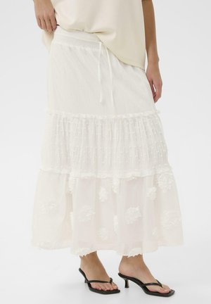 Žena u bijeloj slojevitnoj suknji s cvjetnim uzorcima, uparena s crnim sandalama na petu, stoji ispred jednobojne pozadine.