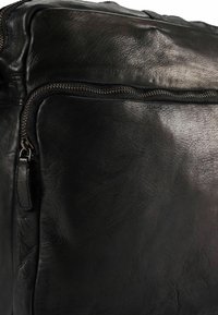 Borsa in pelle nera con superficie texturizzata, cuciture evidenziate e una tasca con zip visibile con dettagli in metallo bronzato.