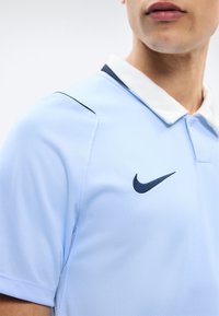 Mann trägt ein hellblaues Nike-Poloshirt mit weißem Kragen und dunkelblauem Logo, gezeigt vom Hals bis zur Mitte des Oberkörpers.