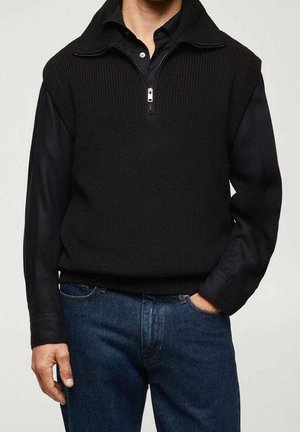 Mężczyzna ubrany w czarną dzianinową kamizelkę bez rękawów z krótkim zamkiem błyskawicznym na czarnej koszuli z długim rękawem i niebieskich dżinsach, z jedną ręką w kieszeni.