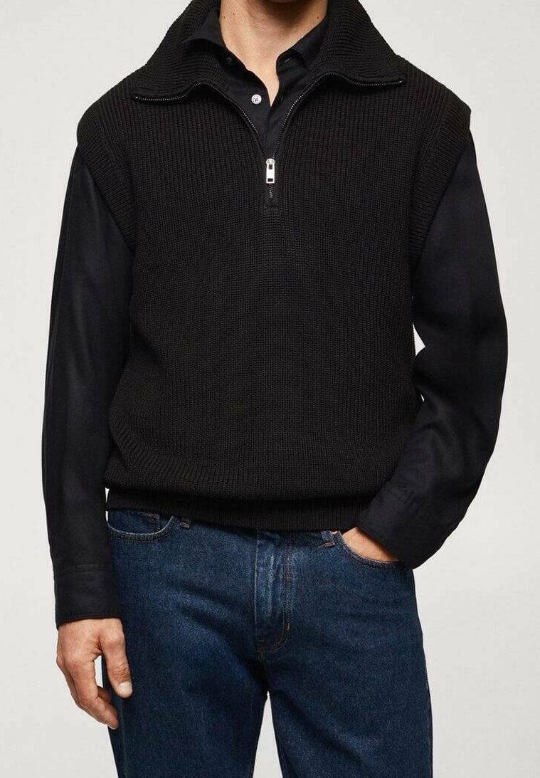 Homme portant un pull sans manches en maille noire avec une courte fermeture éclair par-dessus une chemise noire à manches longues et un jean bleu, une main dans la poche.