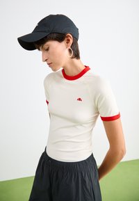 T-shirt crème ajustée à manches courtes avec des poignets et un col rouge côtelé ; petit logo sur la poitrine. Associée à un short noir et une casquette noire.