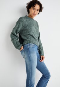 Sweater: textured green knit with cable pattern. Jeans: light blue, fitted with pockets. Model stands sideways against a neutral background.