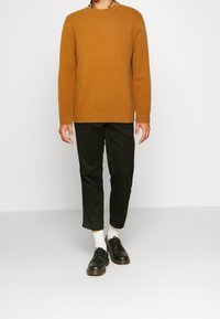Persona che indossa un maglione senape, pantaloni neri a vita alta, calzini bianchi e scarpe nere con lacci su uno sfondo chiaro uniforme.