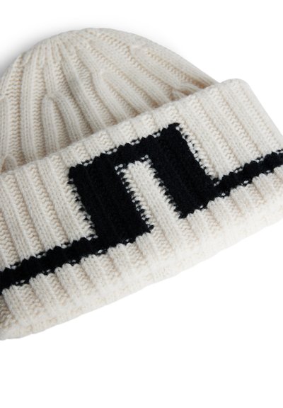 Kreem värvi kudumisbeanie musta soonikuäärisega, millel on julge logodesain. Tekstuur on pehme ja müts istub tihedalt.