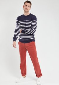 Pull en tricot rayé bleu marine et blanc avec des détails de boutons sur l'épaule, associé à un pantalon rouge retroussé et des baskets blanches.