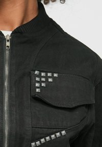 Črna bombažna jakna z rebrastim ovratnikom, zadrgo in srebrnimi kovinskimi dodatki na žepu in rami za kontrast v teksturi.