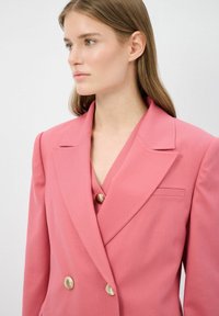 Vaaleanpunainen kaksirivinen bleiseri, jossa on terävät läpät, kullansävyiset napit ja istuva muotoilu, valmistettu jämäkästä kankaasta.