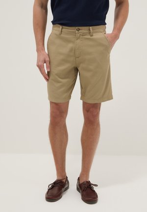Man wearing beige shorts, dark navy shirt, and brown leather lace-up shoes, standing with one hand in pocket.