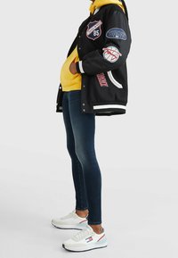Chaqueta universitaria negra con parches, sudadera amarilla debajo, pantalones ajustados azul oscuro y zapatillas blancas con acentos rojos y azules.