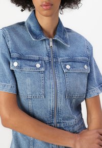 Jeans kombinezon u svijetloplavoj boji, sa zatvaračem sprijeda, kratkim rukavima, dva džepa na prsima i srebrnim dugmadima na pritiskanje s glatkom teksturom.