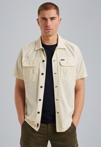 Chemise à manches courtes, couleur beige, avec deux poches poitrine, boutons noirs, et une coupe décontractée, superposée sur une chemise noire.