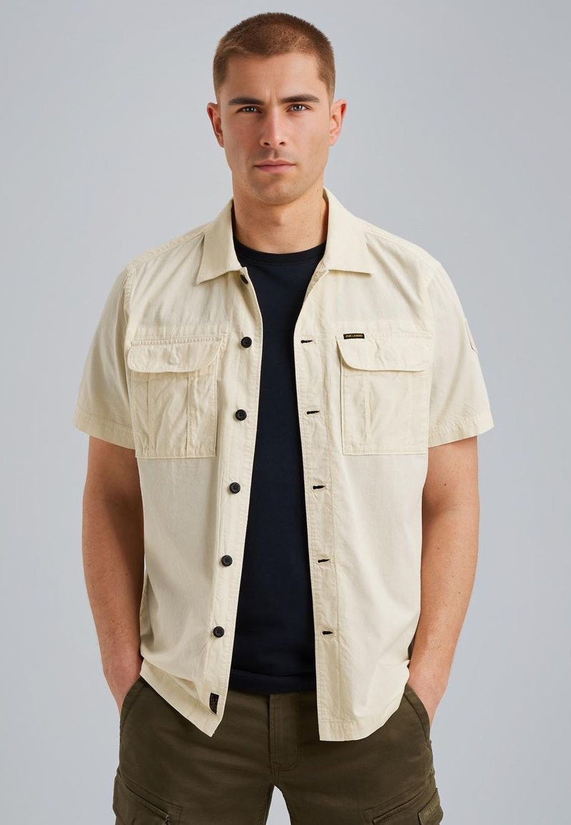 Chemise à manches courtes, couleur beige, avec deux poches poitrine, boutons noirs, et une coupe décontractée, superposée sur une chemise noire.