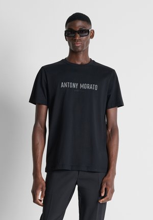 Man wearing black short-sleeve t-shirt with "Antony Morato" text, black pants, and black rectangular sunglasses, standing against a plain background.