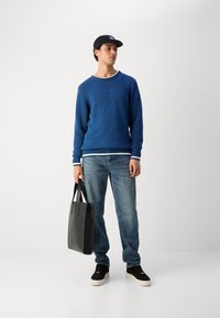 Blå teksturert genser med rund hals, kombinert med lyseblå jeans og svarte sneakers. En svart totebag holdes i den ene hånden.
