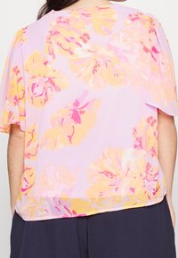 Blusa de color lila claro con mangas cortas que presenta un estampado floral en rosa y amarillo. Textura suave, corte relajado y dobladillo redondeado.