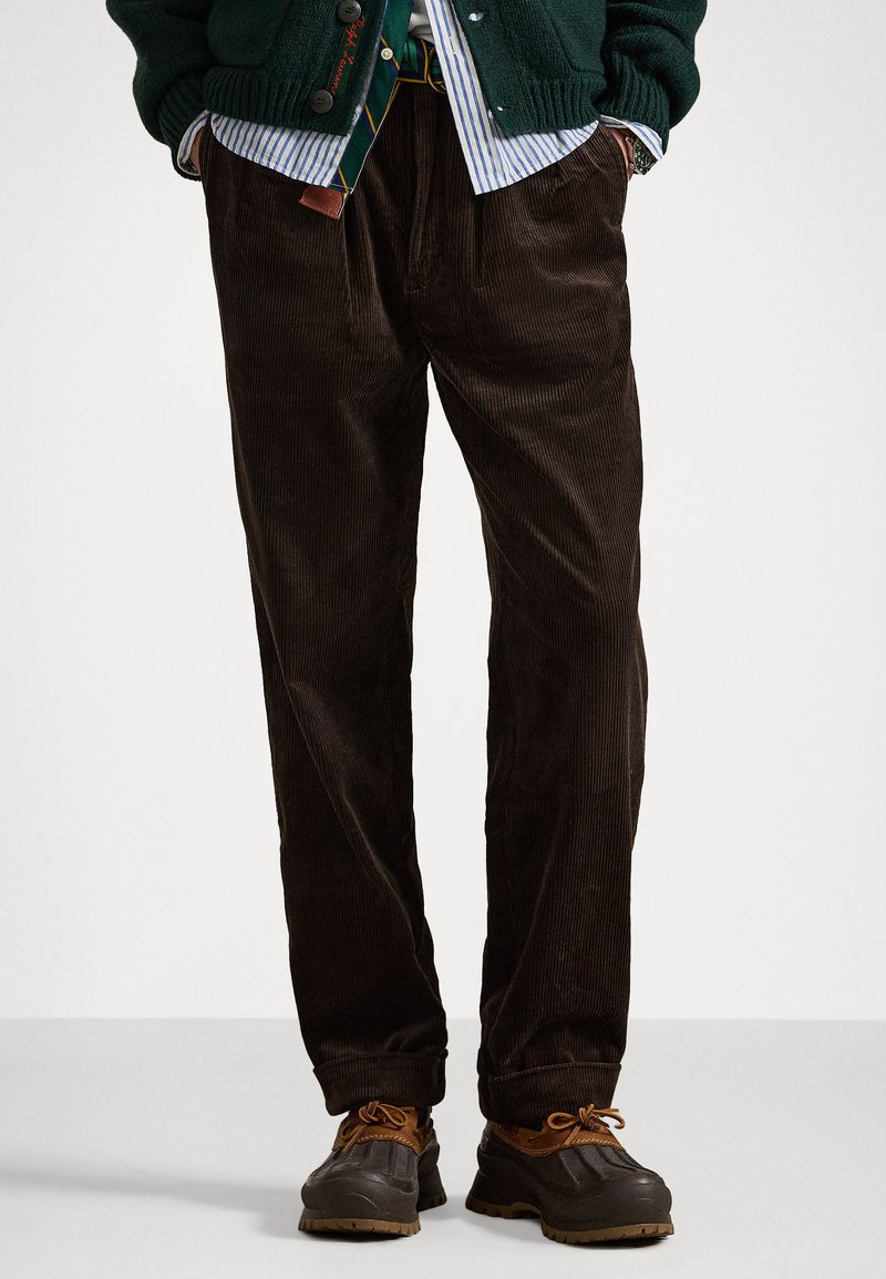 Barna kordbársony nadrág széles lábakkal, függőleges bordázott textúrával. Zöld pulóverrel és barna cipővel viselve.