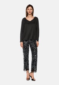 Eine Frau trägt eine schwarze Bluse mit langen Ärmeln und einem breiten Kragen, schwarze Hosen mit Pailletten-Muster, schwarze Ankle-Strap-Absätze und silberne Ohrringe.