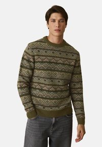 Férfi zöld, barna és bézs cikk-cakk mintás pulóverben és sötétszürke farmernadrágban, egy sima világos háttér előtt áll.