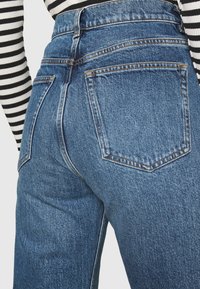 Högmidjade blå denimjeans med en bleknad textur, med två bakfickor och kontrasterande sömmar. Horisontella ränder synliga ovanför.