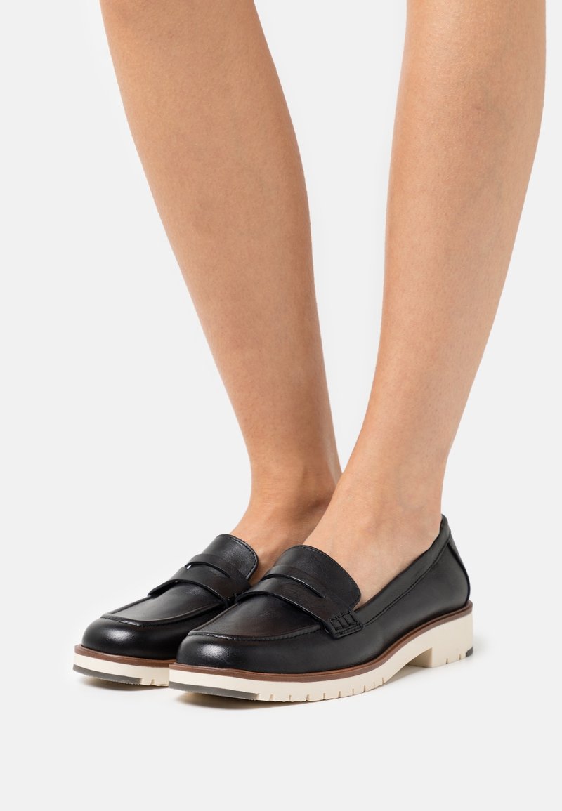 Mocassins en cuir noir avec une bride rembourrée, dotés d'une semelle blanche épaisse et d'un accent marron. Texture lisse et design épuré.