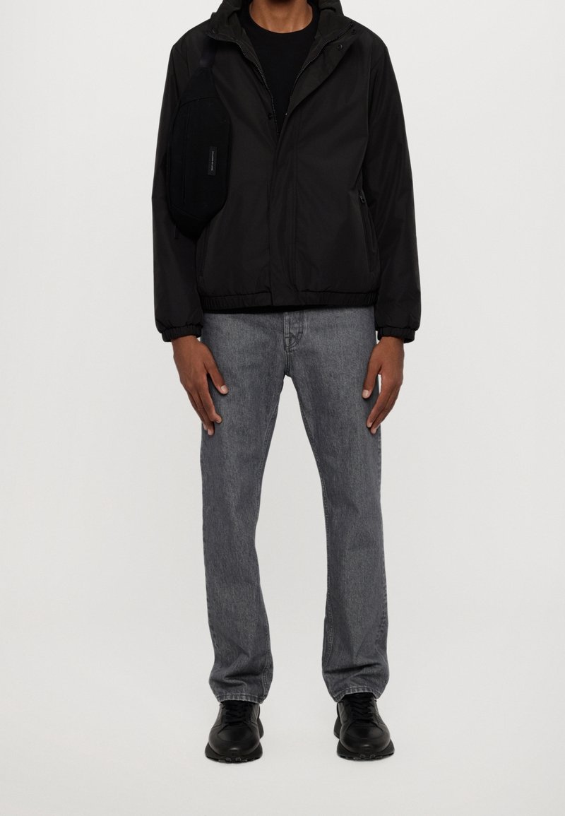 Černá bunda na zip s kapucí, nošená přes černé tričko; světle šedé džínové kalhoty; černé tenisky; malá černá kabelka přes rameno.