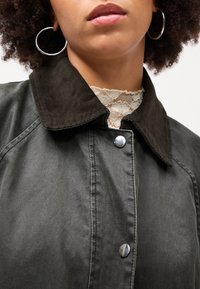 Pilkas švarkas su tamsiu kordūriniu apykakle, sidabriniu spaudimo mygtuku ir tekstūruotu, lygiu audiniu. Kaklo srityje matomas sluoksniuotas nėrinių detalės.