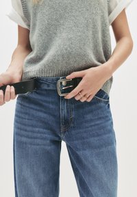 Osoba zapínajúca čierny kožený opasok na modrých džínoch, oblečená v šedom pletenom svetri overene bielej košeli s krátkym rukávom, a so zlatými prsteňmi na pravej ruke.