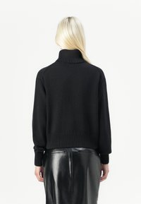 Zwarte gebreide coltrui met geribbelde manchetten en onderzoom, gedragen over een glanzende zwarte leren rok. Achteraanzicht toont een zachte textuur en een ontspannen pasvorm.
