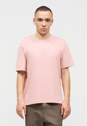 Νεαρό άτομο με κοντά μαλλιά που φοράει ένα απλό ανοιχτό ροζ κοντομάνικο μπλουζάκι και καφέ παντελόνι, στέκεται μπροστά σε ένα λευκό φόντο.