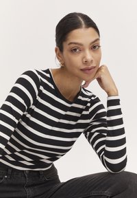 Gestreiftes Langarmhemd in Schwarz und Weiß aus weicher Stoffqualität, kombiniert mit dunklen Jeans. Das Model stützt das Kinn auf die Hand gegen einen schlichten Hintergrund.