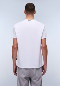 Camiseta blanca de algodón con mangas cortas, cuello redondo y un sutil detalle de etiqueta en la parte inferior izquierda. Textura suave y corte relajado.
