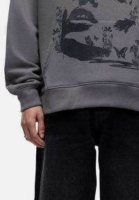 Felpa grigia con una stampa grafica di labbra, farfalle e fiori. Abbinata a jeans neri, che mostrano una vestibilità casual e rilassata.