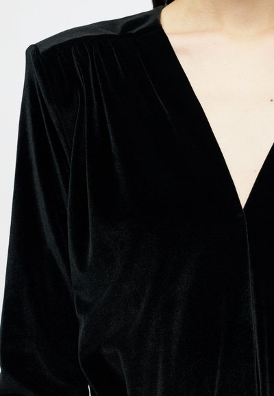 Top din catifea neagră cu mâneci lungi și decolteu adânc în V, purtat de o persoană cu pielea deschisă, arătată din umeri până la mijlocul trunchiului.