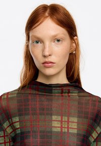Przezroczysta bluzka w kratę z zielonymi, czerwonymi i czarnymi wzorami. Modelka ma długie rude włosy i nosi prosty złoty kolczyk. Czysta skóra.