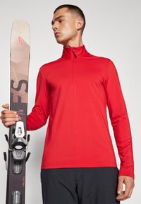 Haut à manches longues rouge avec un col zippé mi-hauteur, texture lisse et design ajusté. Un skieur tient des skis noir et blanc avec des fixations visibles.