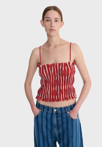 Jeune femme portant un haut sans manches froncé à rayures rouges et blanches et un jean taille haute à rayures bleues, les mains dans les poches.