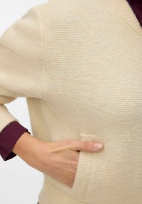 Bēšas adītas džemperis ar V veida kaklu un priekšējo kabatu. Iezīmējas ar ribotiem aproces rokturiem burgundijas krāsā. Mīksta tekstūra un maiga svītru raksts.