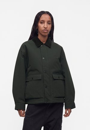 Person med langt flet håret iført en mørkegrøn jakke med knapper, sort krave og store forlommer mod en ensfarvet baggrund.