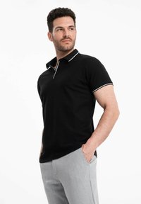 Polo noir avec une accent blanc le long du col et des manches, fabriqué dans un tissu lisse, accompagné d'un pantalon gris clair.