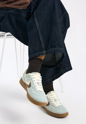 Light blue suede sneakers with white laces and brown accents, paired with dark denim pants featuring a wide leg and visible stitching.